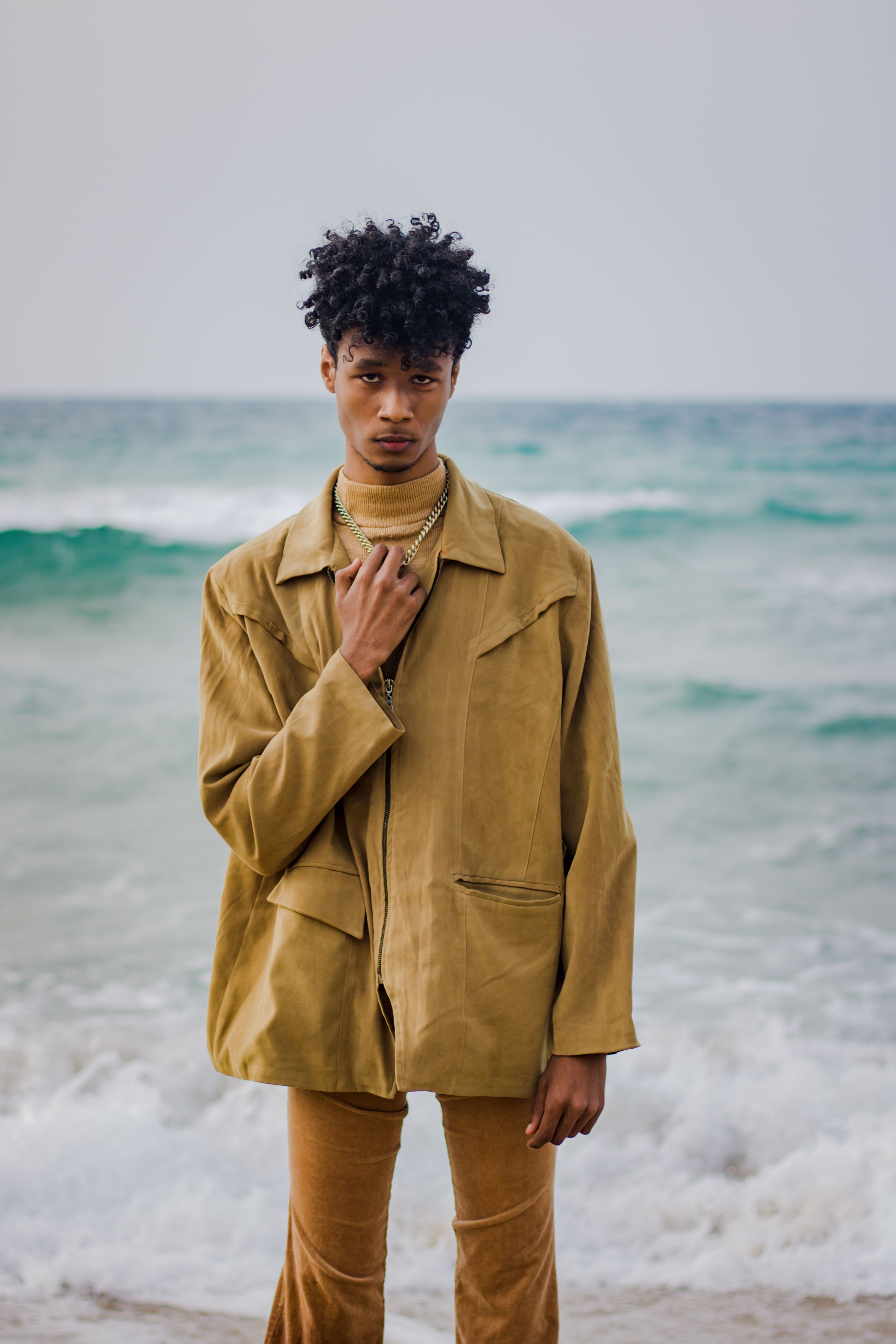 Fashionable young man with curly black hair posing by the sea in a beige outfit, showcasing modern style.