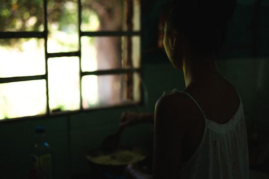 A woman stands by a window, cooking inside a dimly lit room, creating a serene mood.