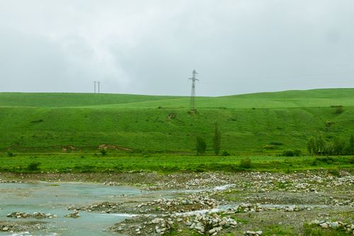 Free Scenic view of lush green hills, a flowing river, and power lines under a cloudy sky. Stock Photo