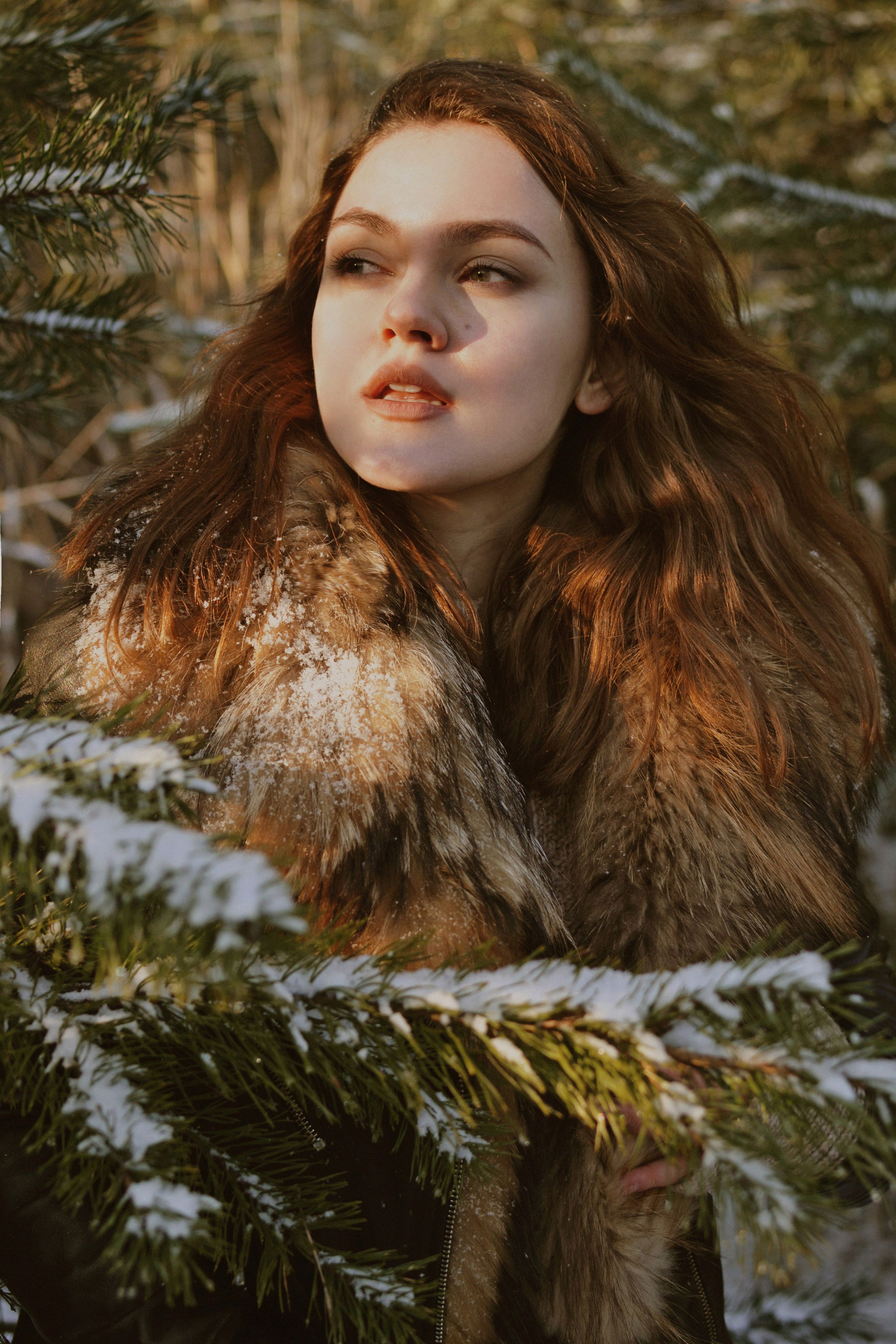 A woman in a fur jacket surrounded by snowy green branches, gazing into the distance.