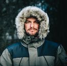 A bearded man wearing a fur-lined parka stands outdoors, capturing a winter scene in England.