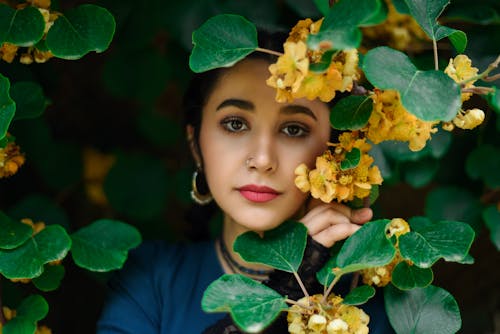 Photo of a Young Woman Standing between Branches with Yellow Flowers and Green Leaves