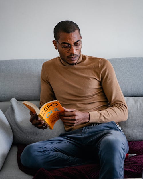 Photo of a Black Man in Glasses Sitting on a Couch and Reading a Book