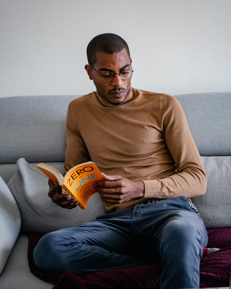 Photo Of A Black Man In Glasses Sitting On A Couch And Reading A Book