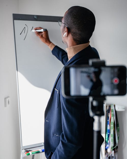 Photo of a Black Man Being Recorded by a Phone while Writing on a Whiteboard