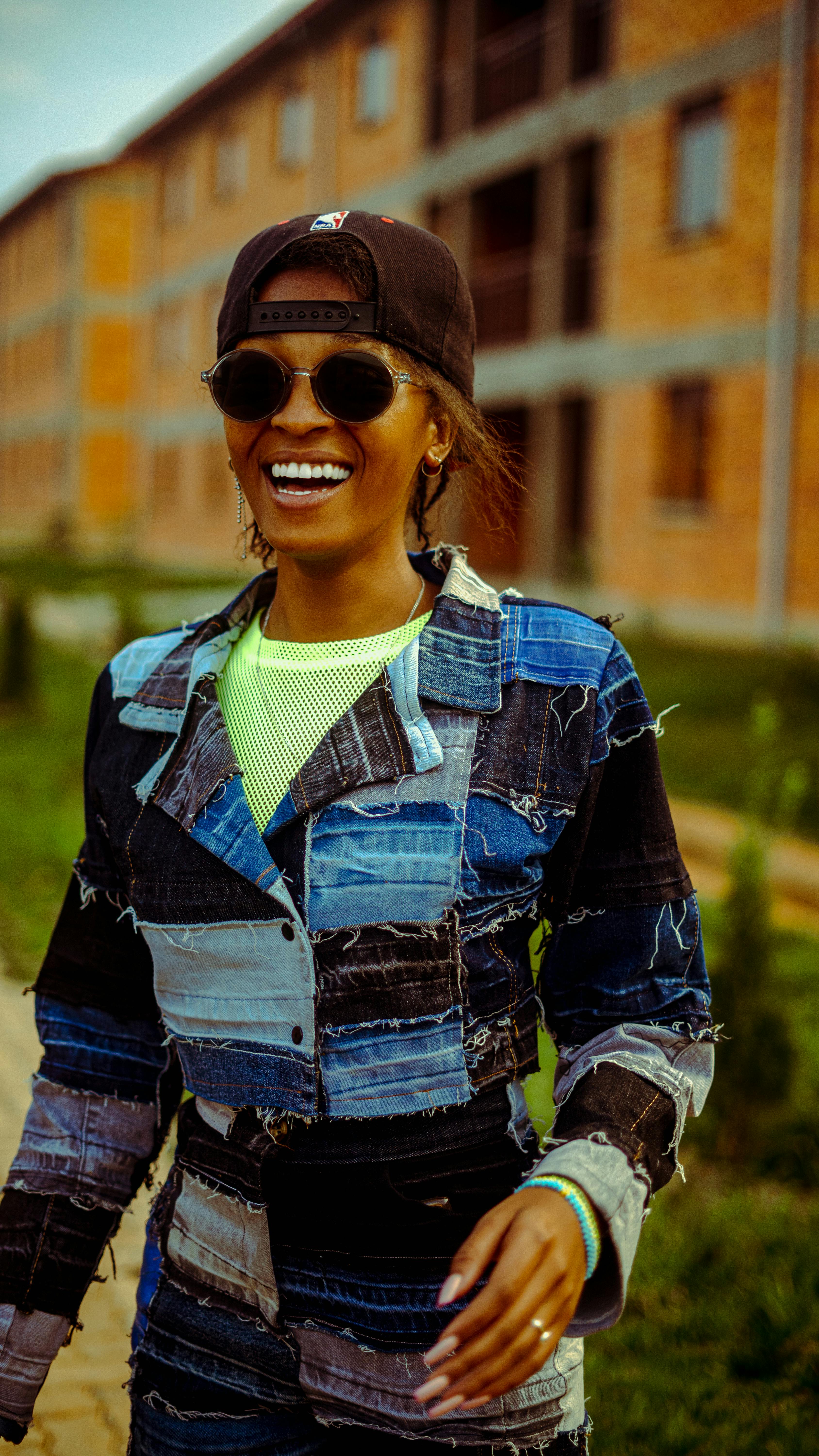 Stylish woman in patchwork denim, sunglasses, and cap, smiling outdoors in Kigali, Rwanda.