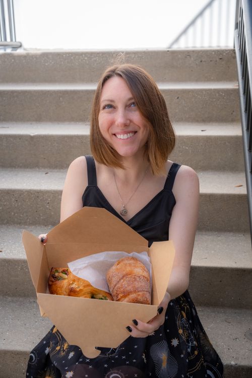 Free Woman sitting on steps with a box of fresh croissants, smiling brightly at the camera. Stock Photo