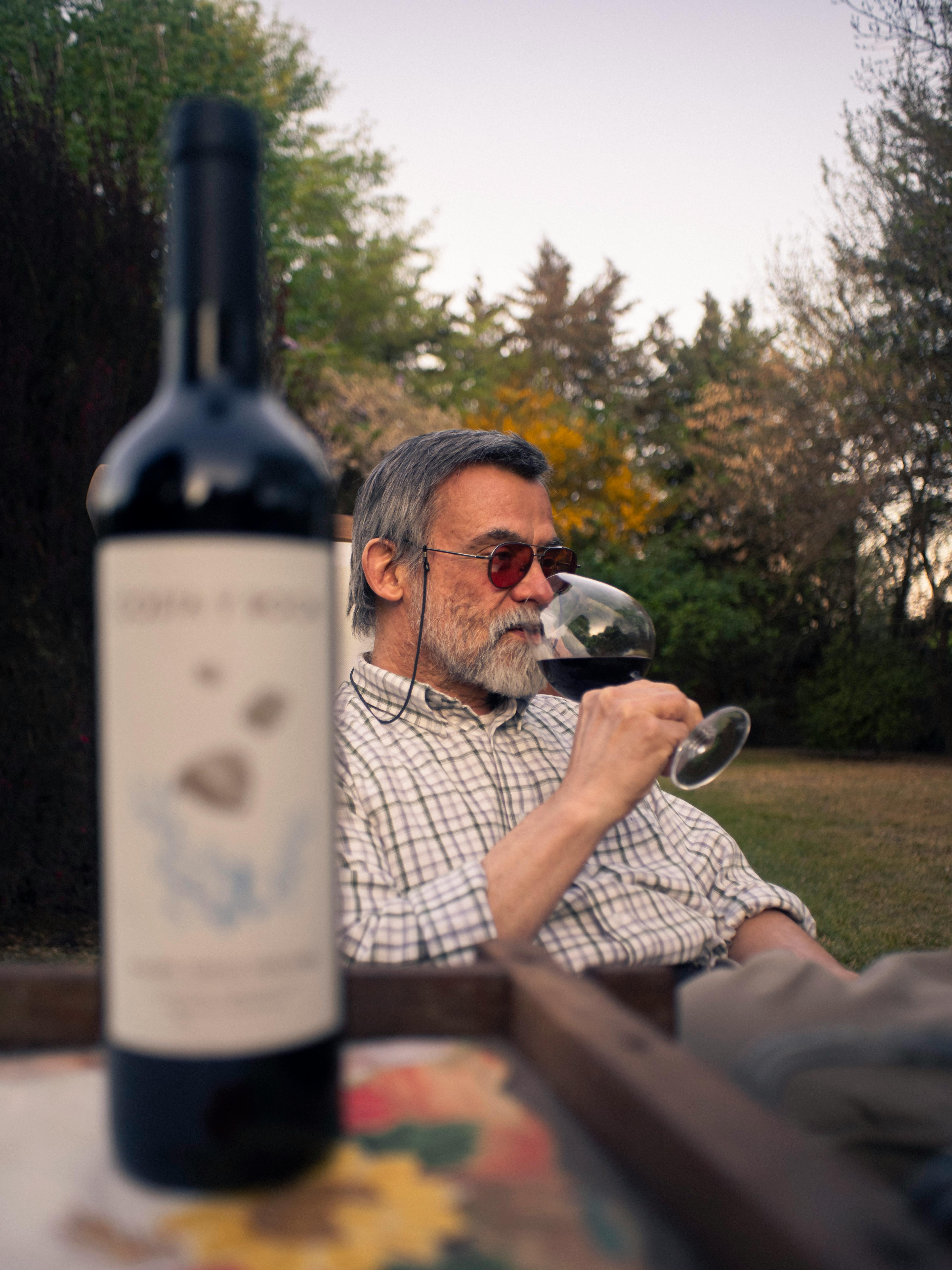 Senior man with sunglasses drinks wine in a serene garden setting with a wine bottle in focus.