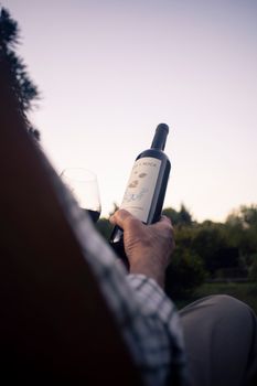 A person holding a wine bottle and glass outside in a tranquil setting.