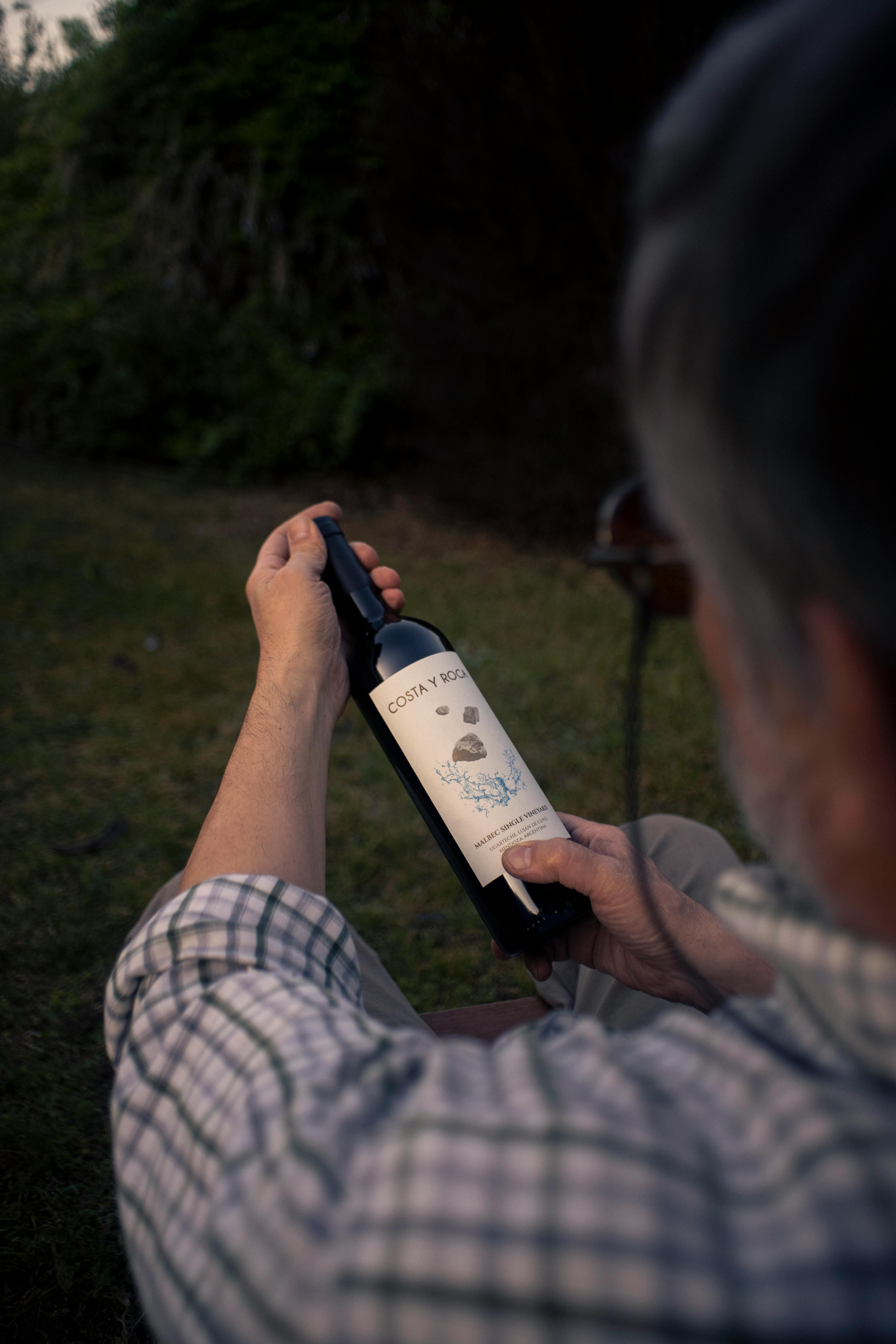 Free A man outdoors at twilight examining the label of a red wine bottle. Stock Photo