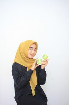 Asian woman smiling, holding beauty products with a headscarf against a white background. - Fotografer Sidoarjo