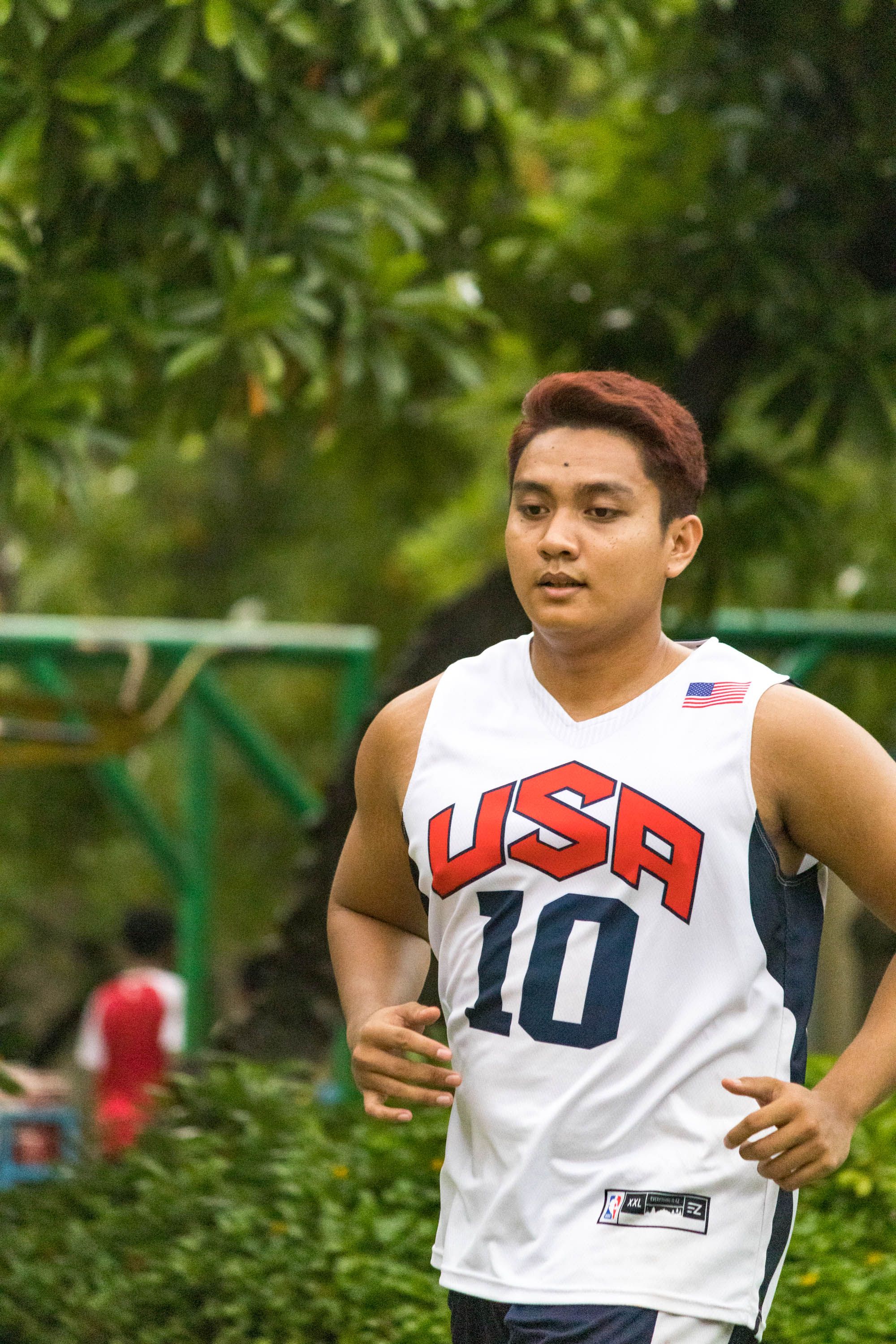Asian man jogging in a park wearing a 'USA' jersey, embracing fitness and healthy lifestyle.