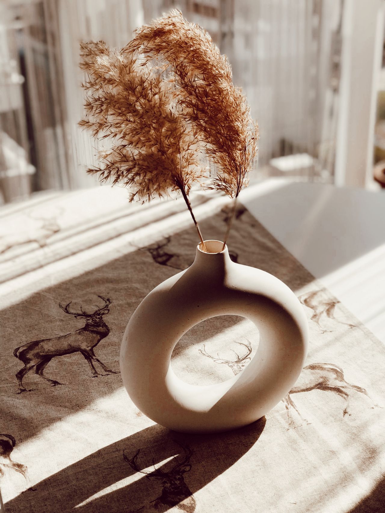 Two dried pampas stems in a round donut-shaped white vase on a deer-print table runner