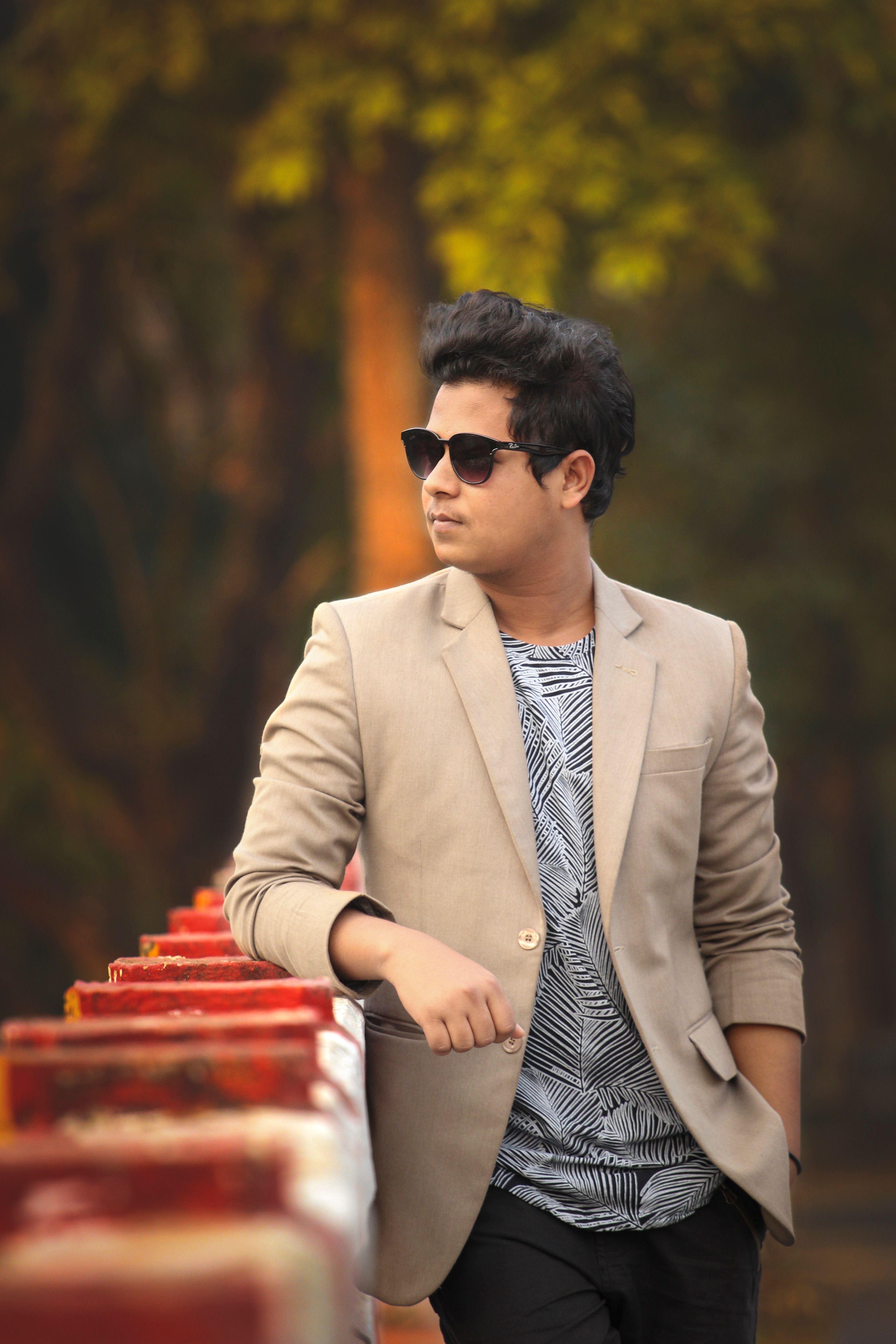 Asian man wearing a blazer and sunglasses, leaning on a railing outdoors. Fashionable and confident.