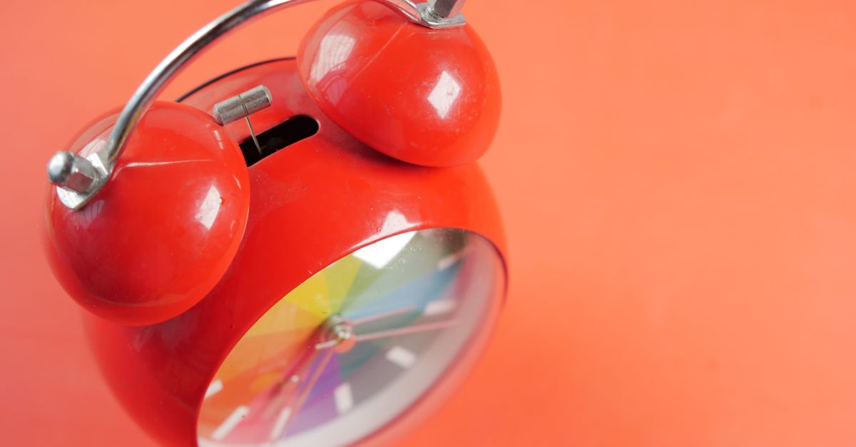 Close-up de um relógio despertador analógico vermelho com um mostrador de arco-íris em um fundo laranja brilhante.