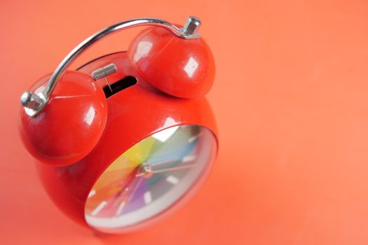 Close-up of a red analog alarm clock with a rainbow clock face on a bright orange background.