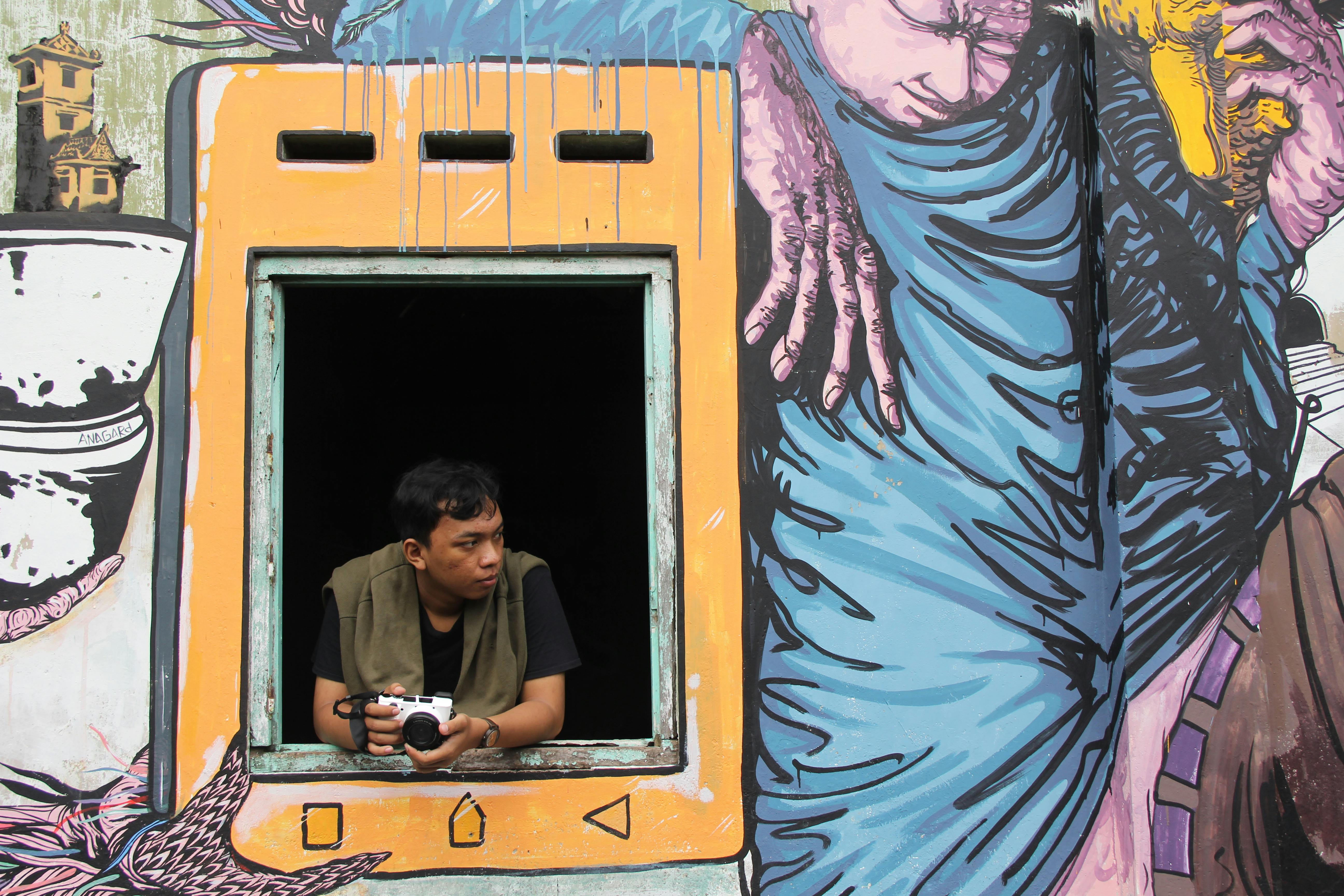 Asian man with camera leaning out window framed by vibrant mural art.