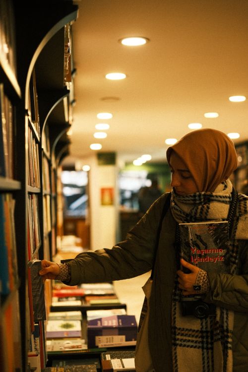 Free Woman in hijab exploring a bookstore aisle, selecting a book with warm lighting. Stock Photo