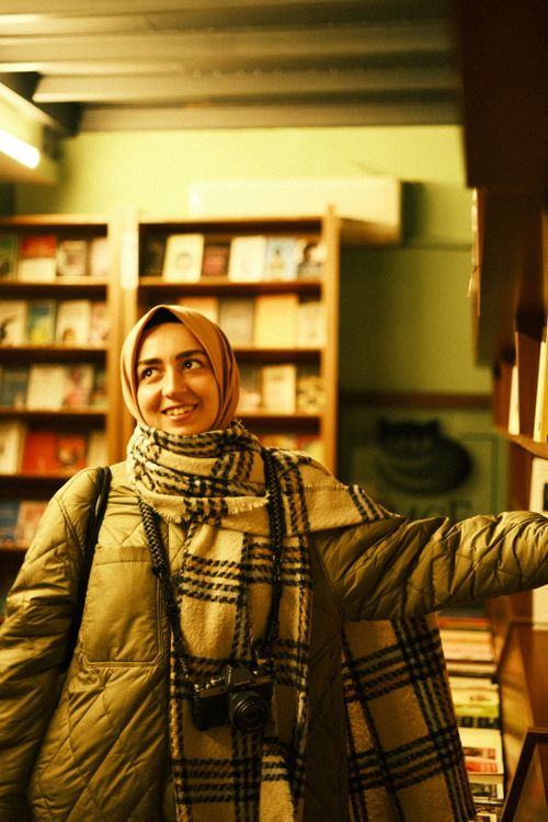 Free Smiling woman in a bookstore with a headscarf and plaid scarf, holding a camera. Warm, inviting scene. Stock Photo