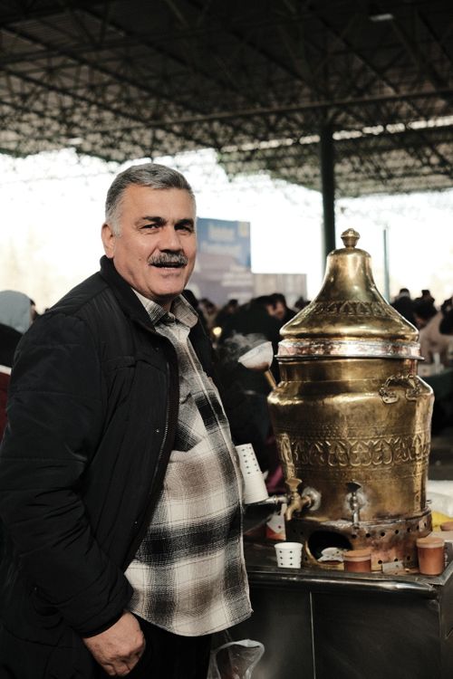 Free Middle-aged man smiling beside a traditional brass samovar at an outdoor market, surrounded by people. Stock Photo