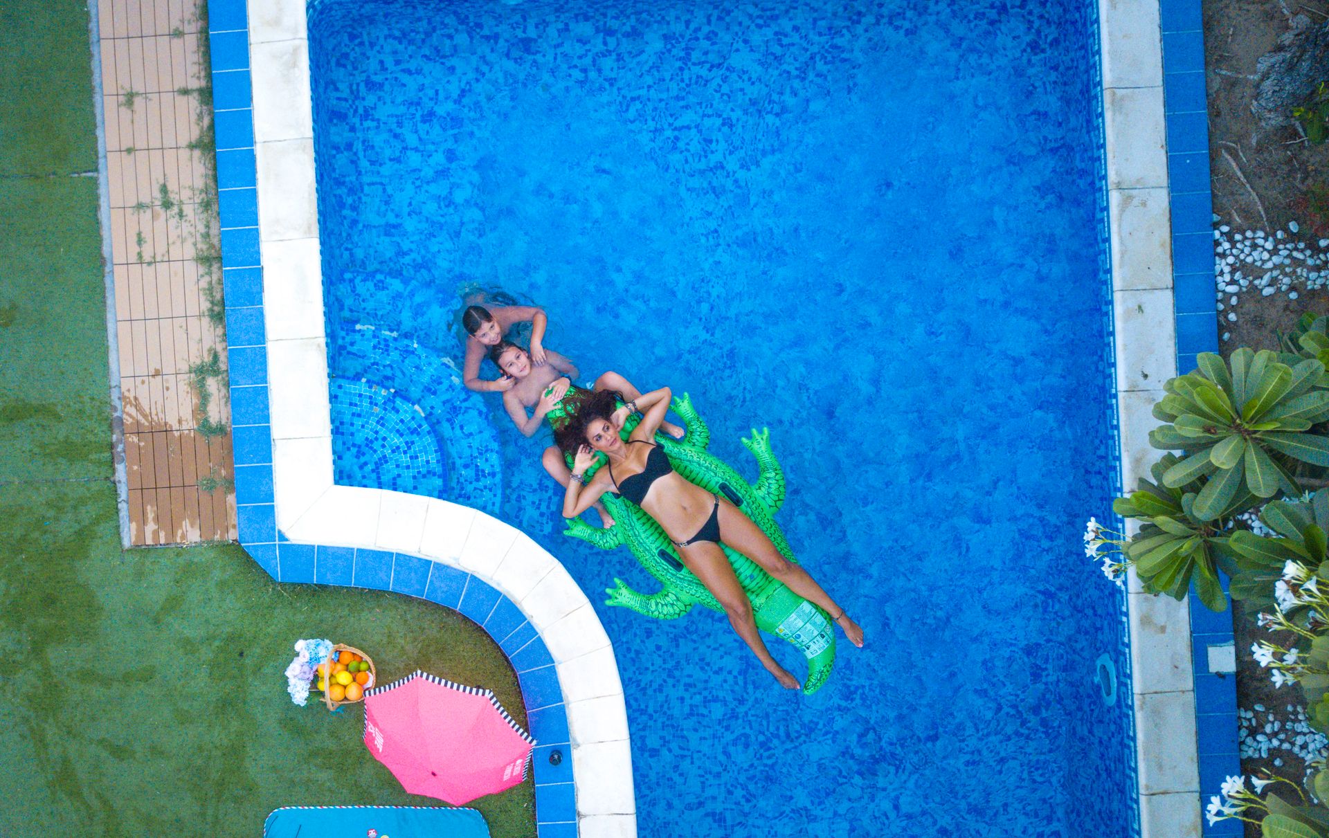 Aerial shot of three people having fun in a pool in Dubai with floaties and refreshments.