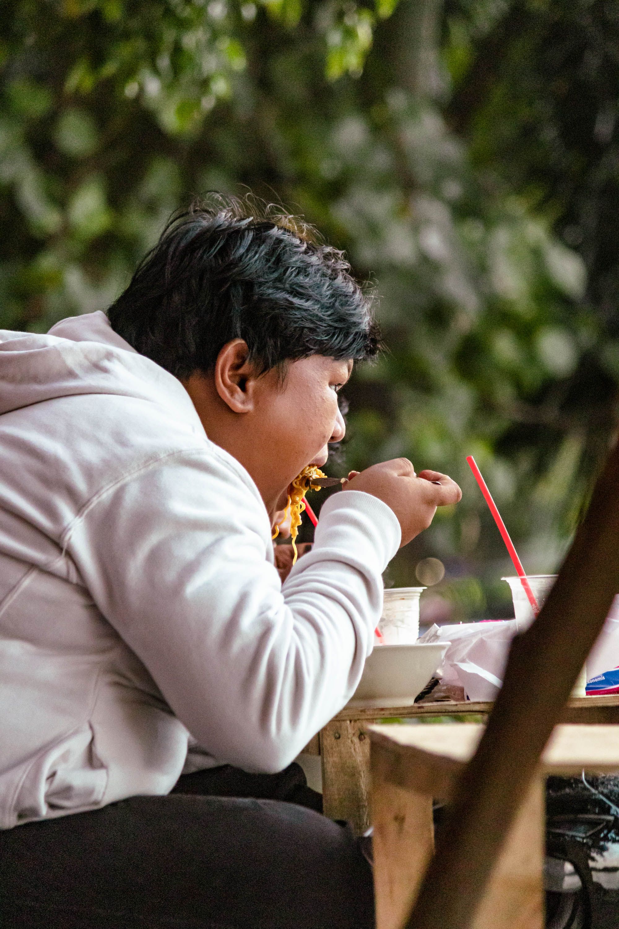 Adult eating noodles at an outdoor table, wearing a white jacket on a street. Captured in a candid moment.
