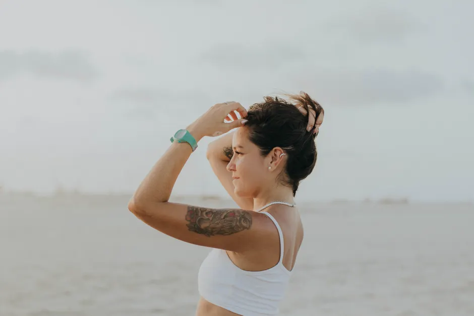 Woman at the beach with a visible arm tattoo, adjusting her hair in natural light