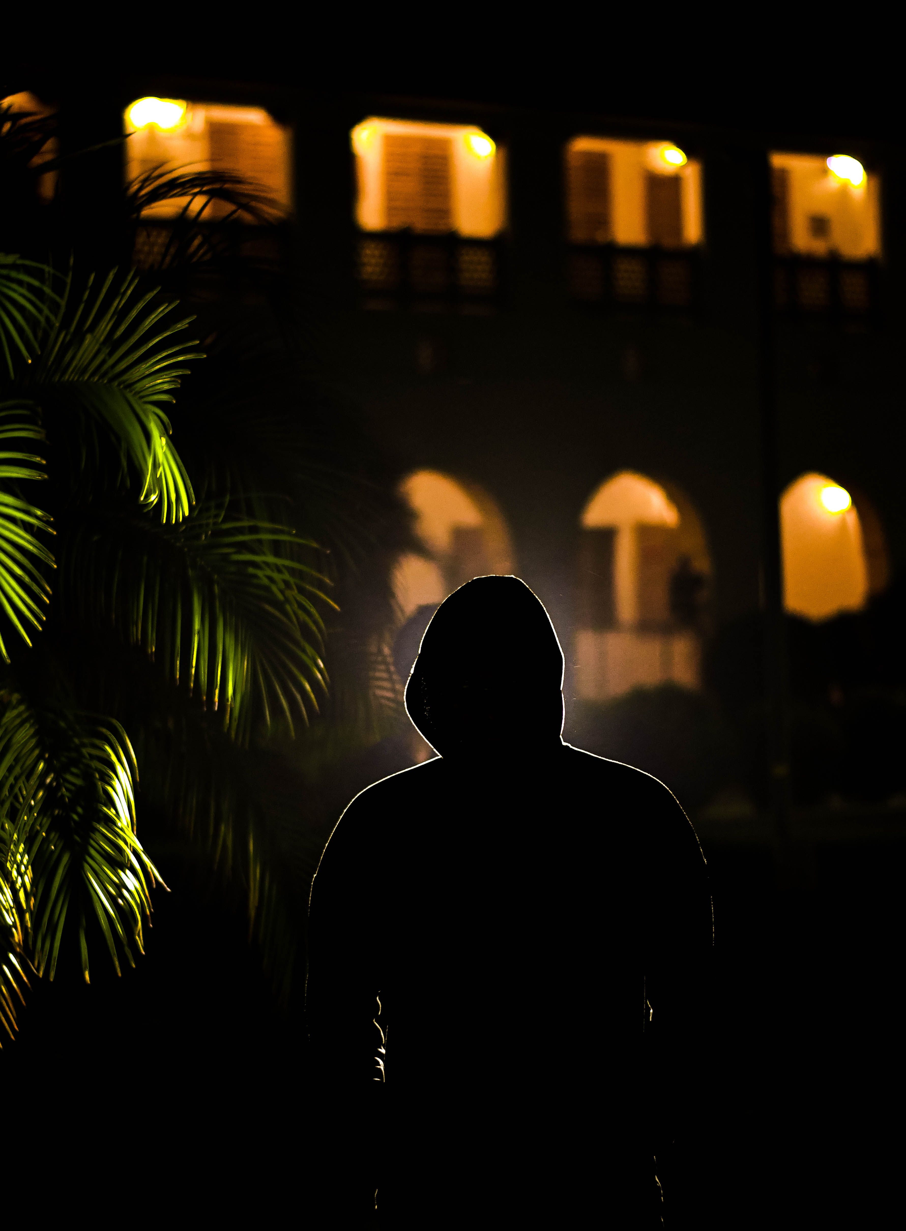 Silhouette of a hooded figure at night in front of a lit building, evoking mystery.