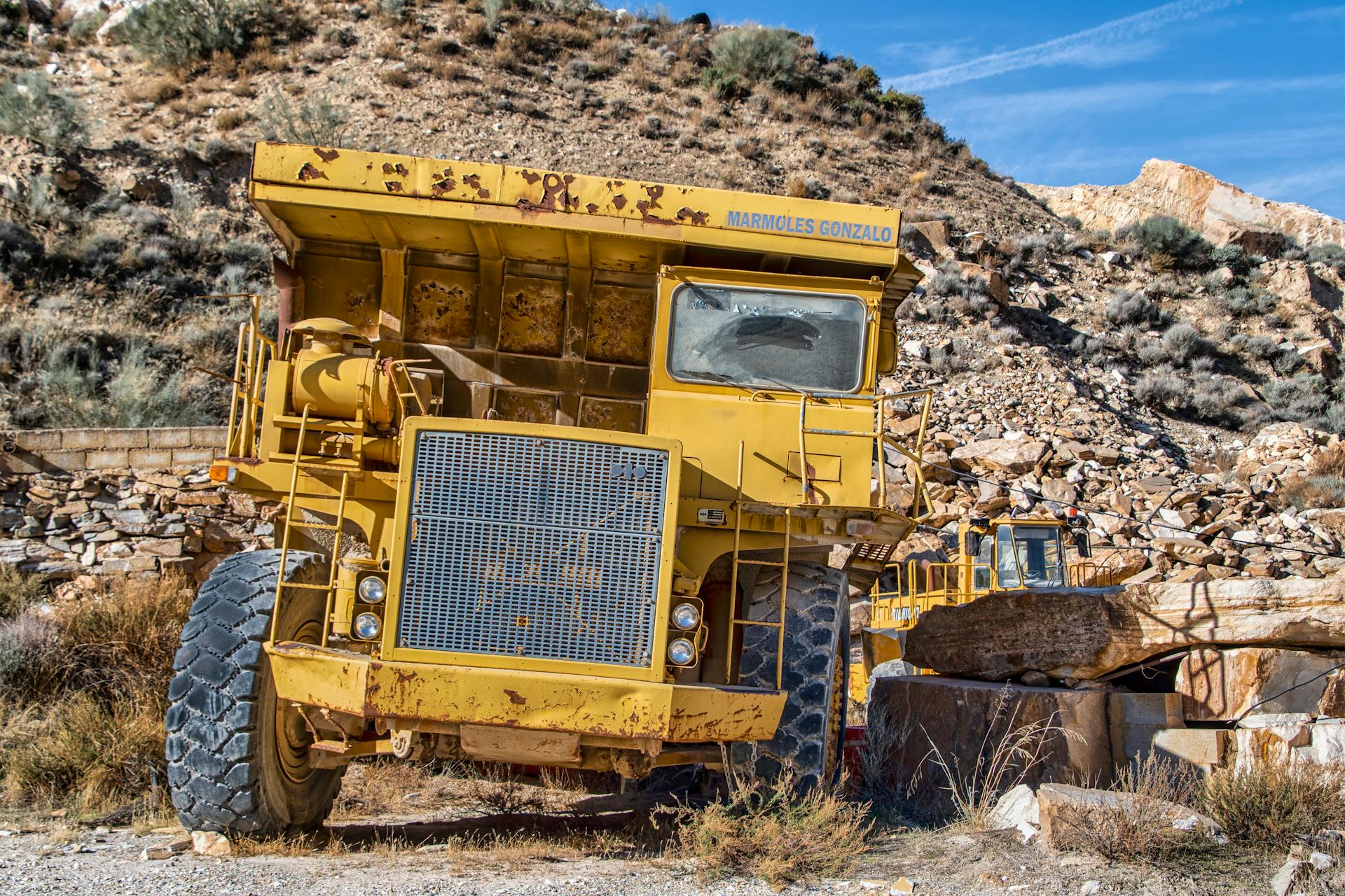 Massive mining truck parked in a rocky, arid quarry with rugged terrain under clear blue skies.