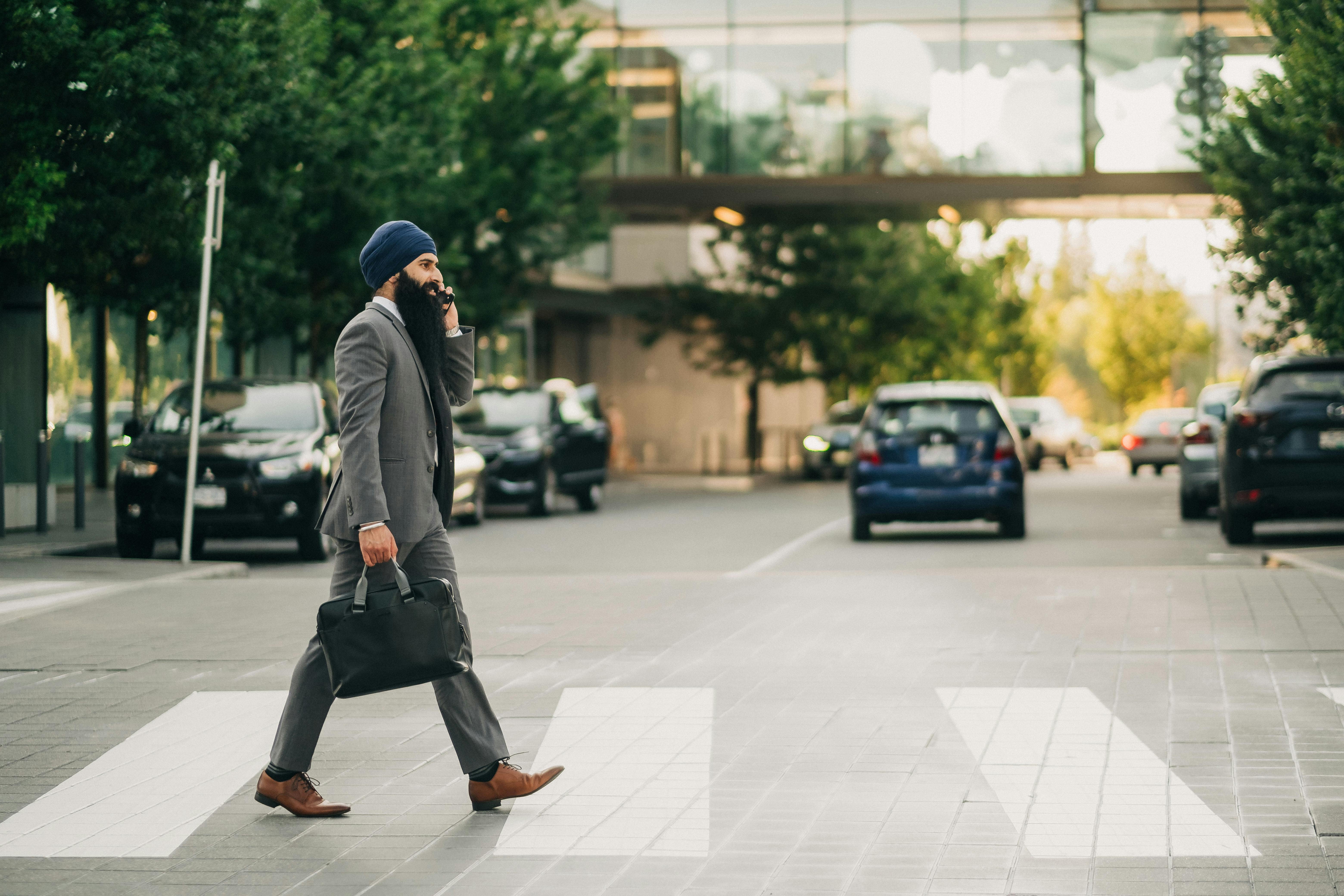 Middle Eastern man in suit and turban walking across city street using phone.