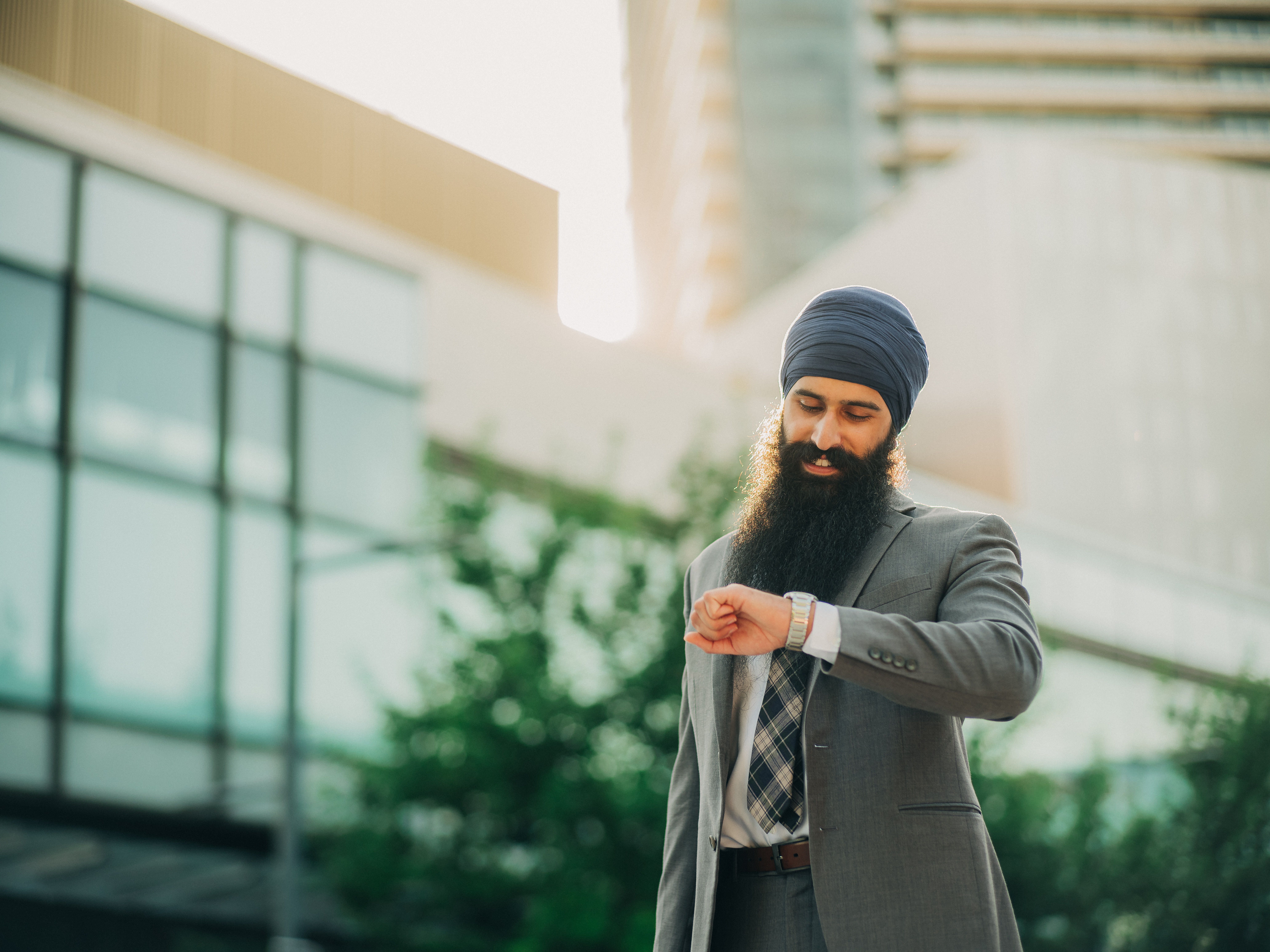A businessman with a turban checks his watch, standing outdoors with modern architecture in the background.