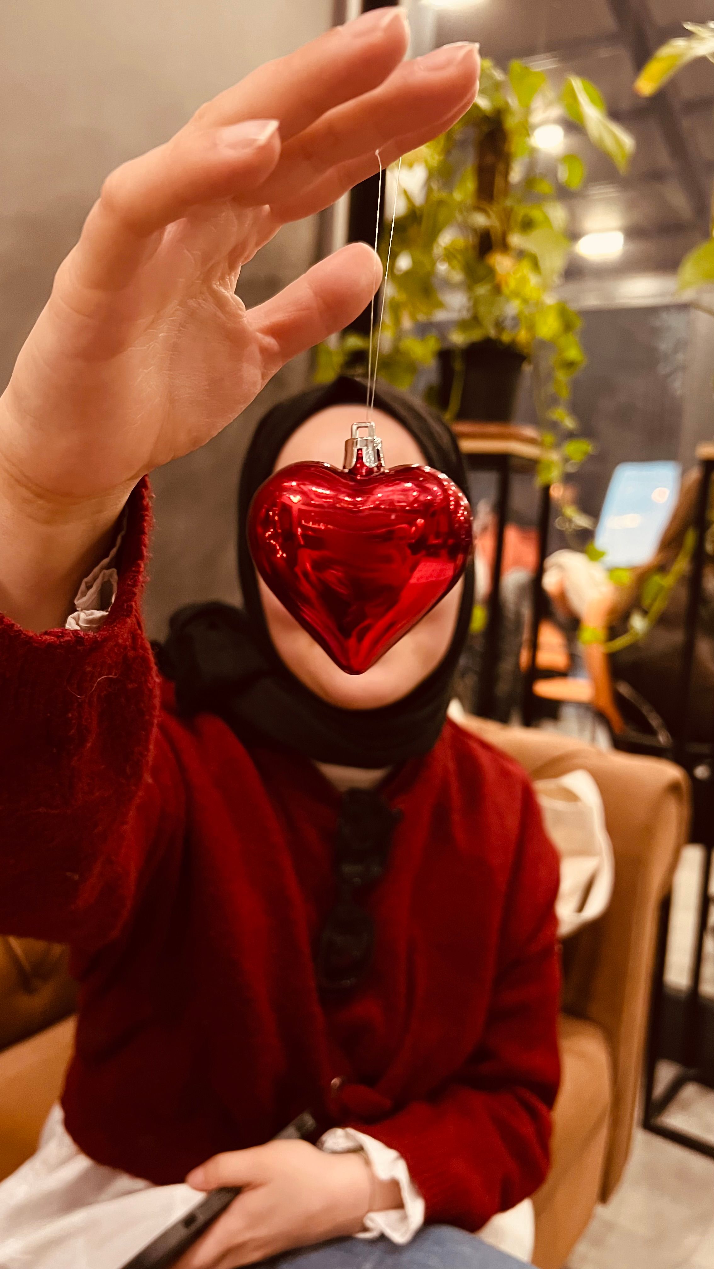 A woman with a red sweater holds a heart-shaped Christmas ornament indoors.