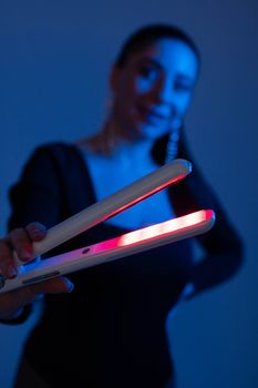A woman holds a glowing hair straightener in a moody blue-lit setting, emphasizing hairstyling tools.