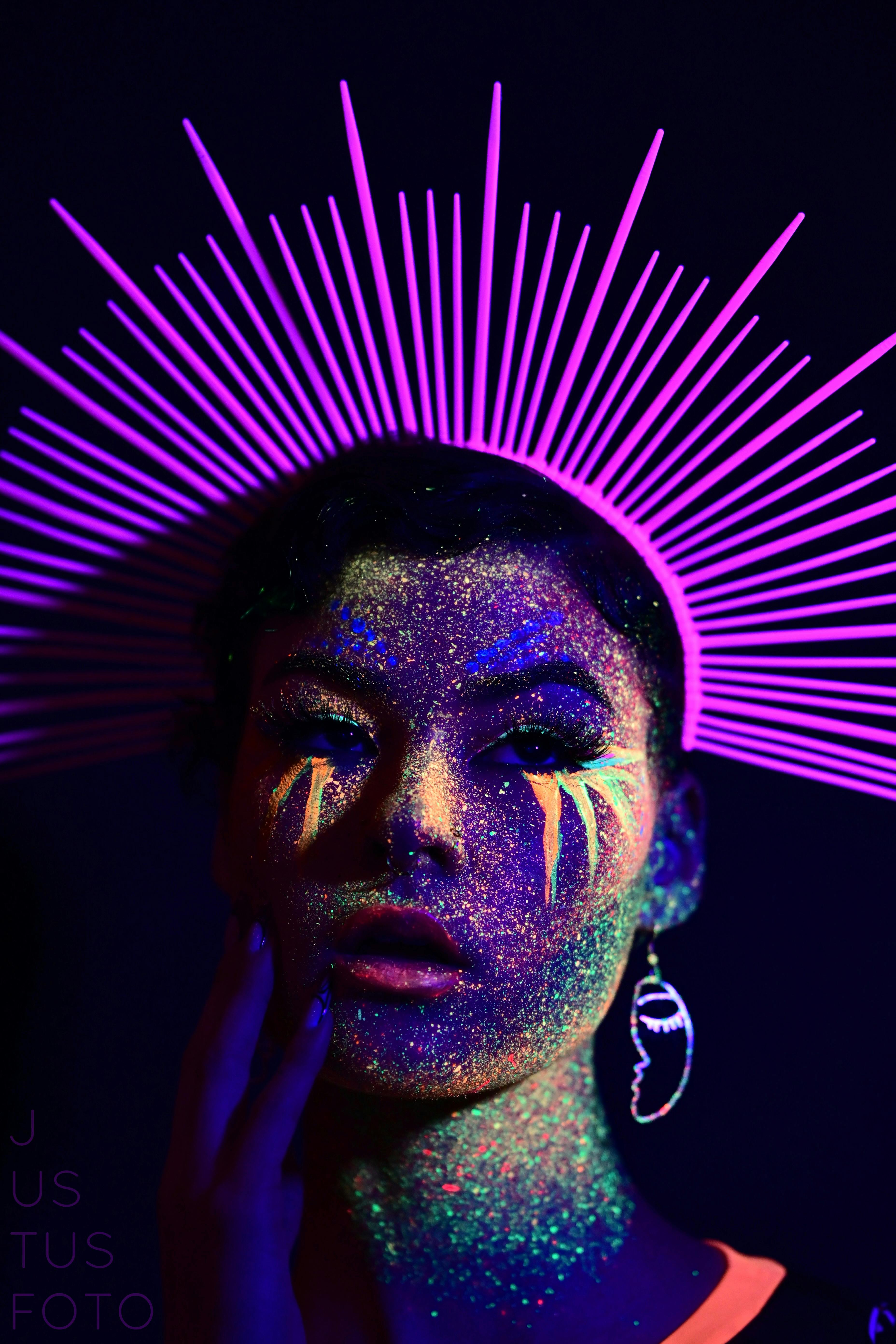Striking portrait of a woman with neon makeup and artistic headpiece under fluorescent lighting.