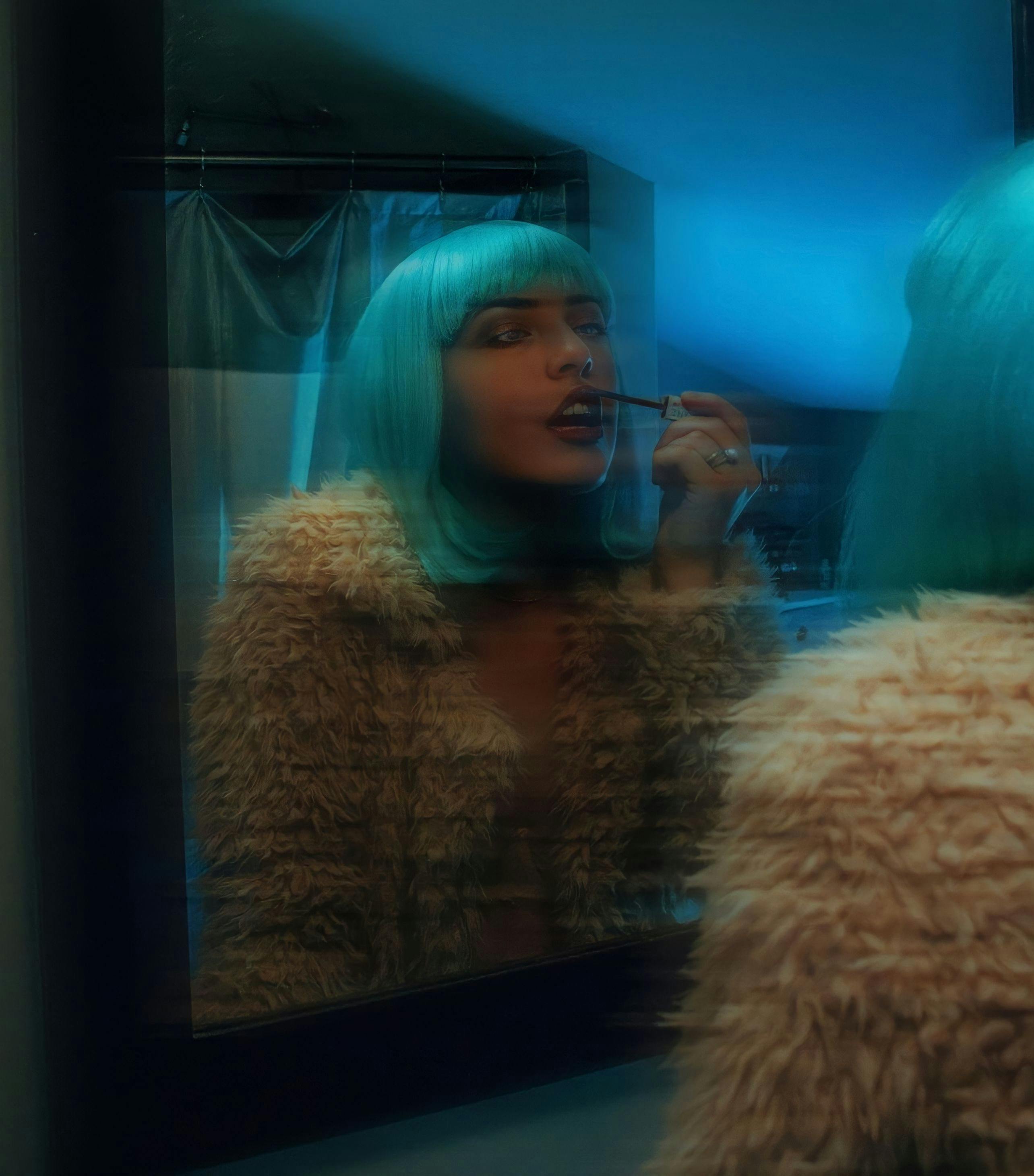 Woman in blue wig and fur coat applying lipstick, seen in mirror reflection.