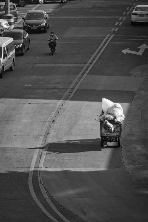 Free A black and white street scene featuring a lone garbage collector pushing a cart amidst city traffic. Stock Photo