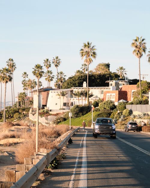 Free Sunlit coastal road lined with palm trees and modern houses, cars driving under a clear sky. Stock Photo