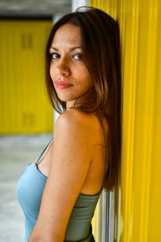 A fashionable woman with long brown hair standing against a bright yellow wall, striking a pose.