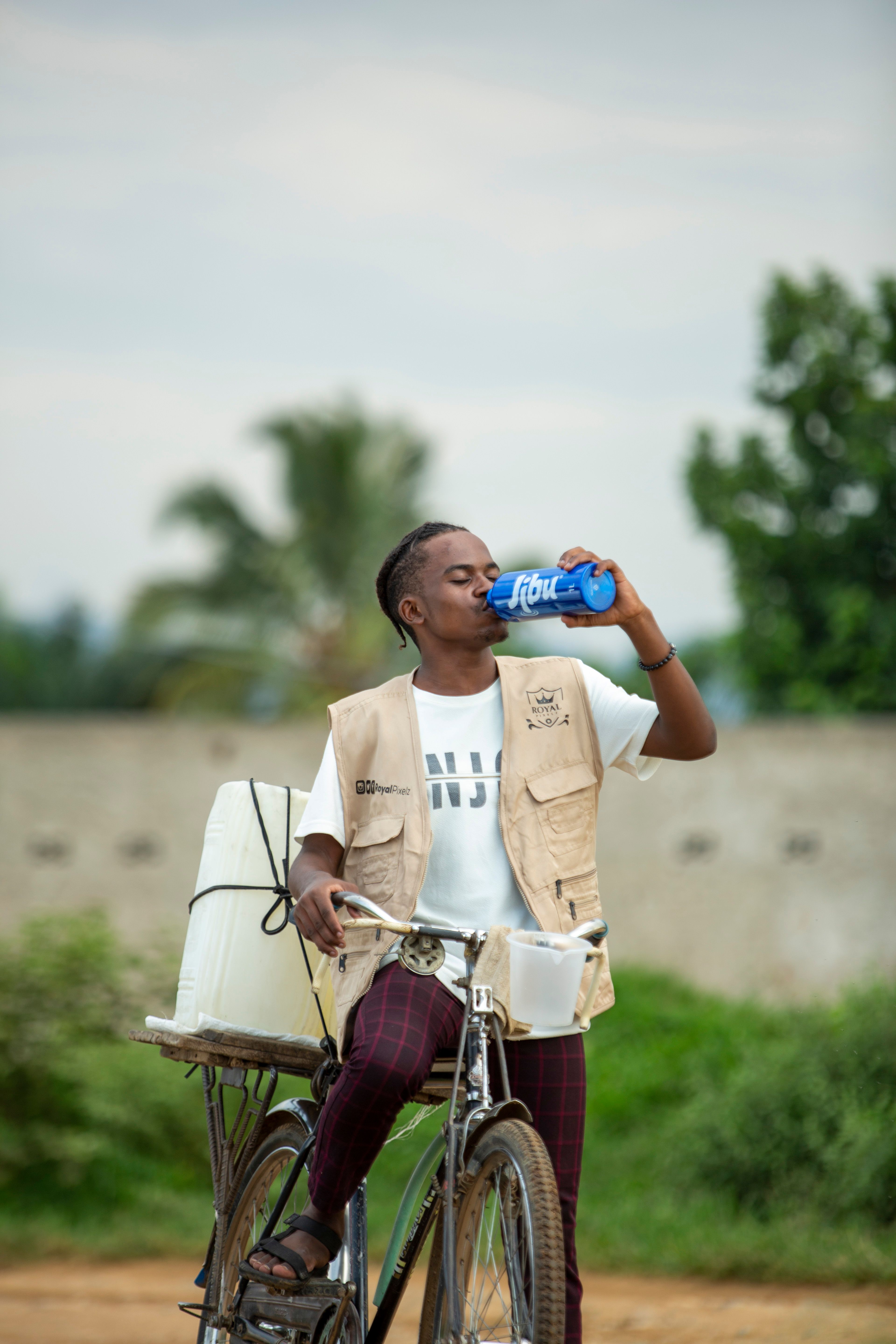 A young man on a bicycle drinking from a bottle in an open outdoor setting.