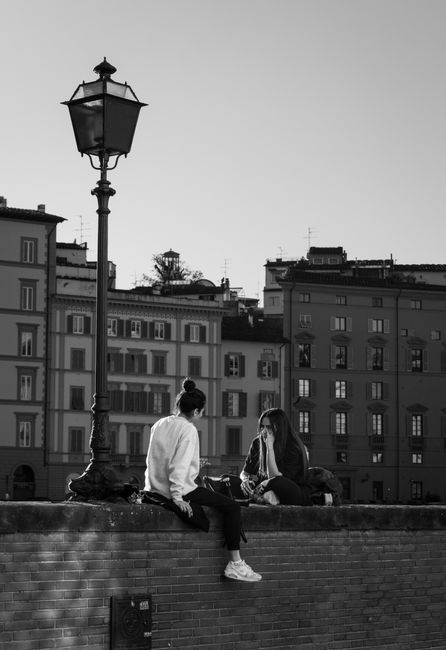 Two women sit on a wall in Tuscany, Italy, with historical buildings in the background.