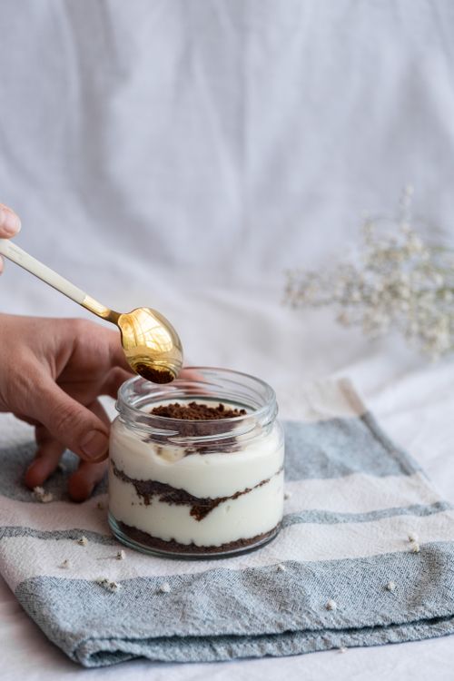 Free A close-up of a hand holding a dessert jar with layers, placed on a cloth with a backdrop of flowers. Stock Photo