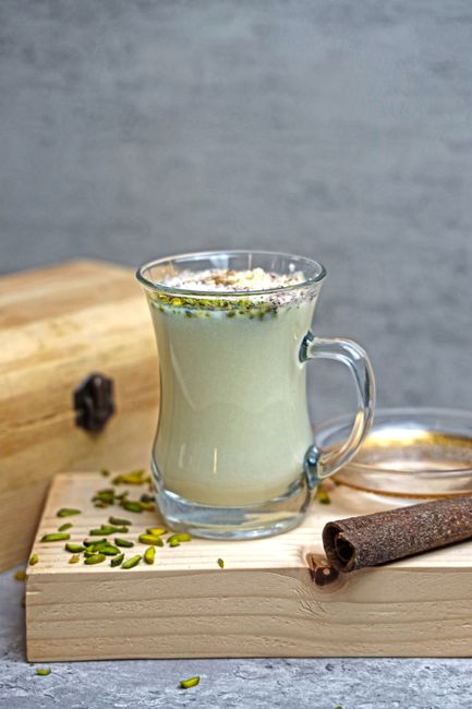 A glass of pale green-tinted pistachio milk next to a small pile of shelled pistachios on a wooden board.
