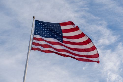 Free Vibrant American flag waving on a flagpole against a bright blue sky with clouds. Stock Photo