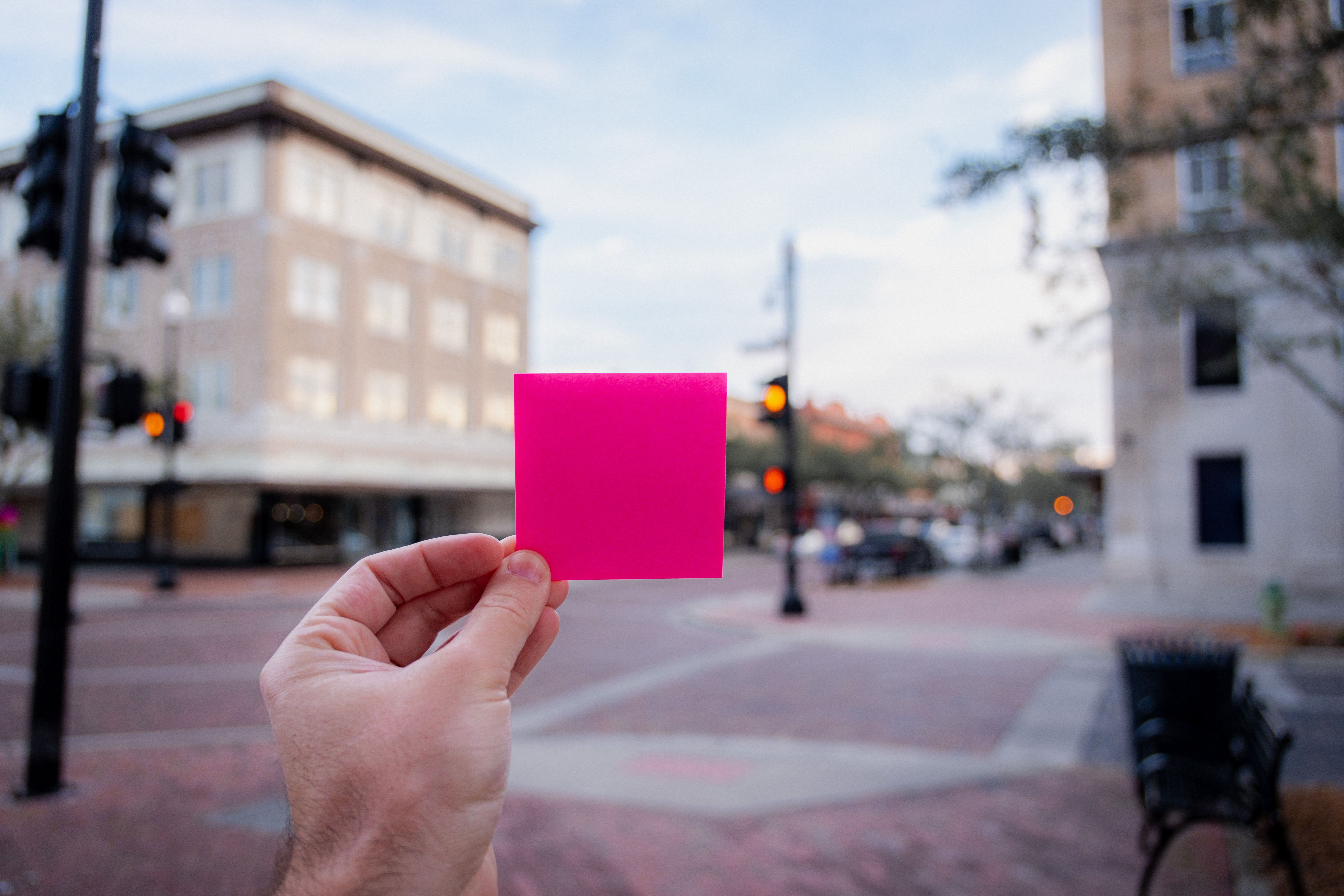 Close-up of a hand with a pink sticky note in an urban outdoor setting.