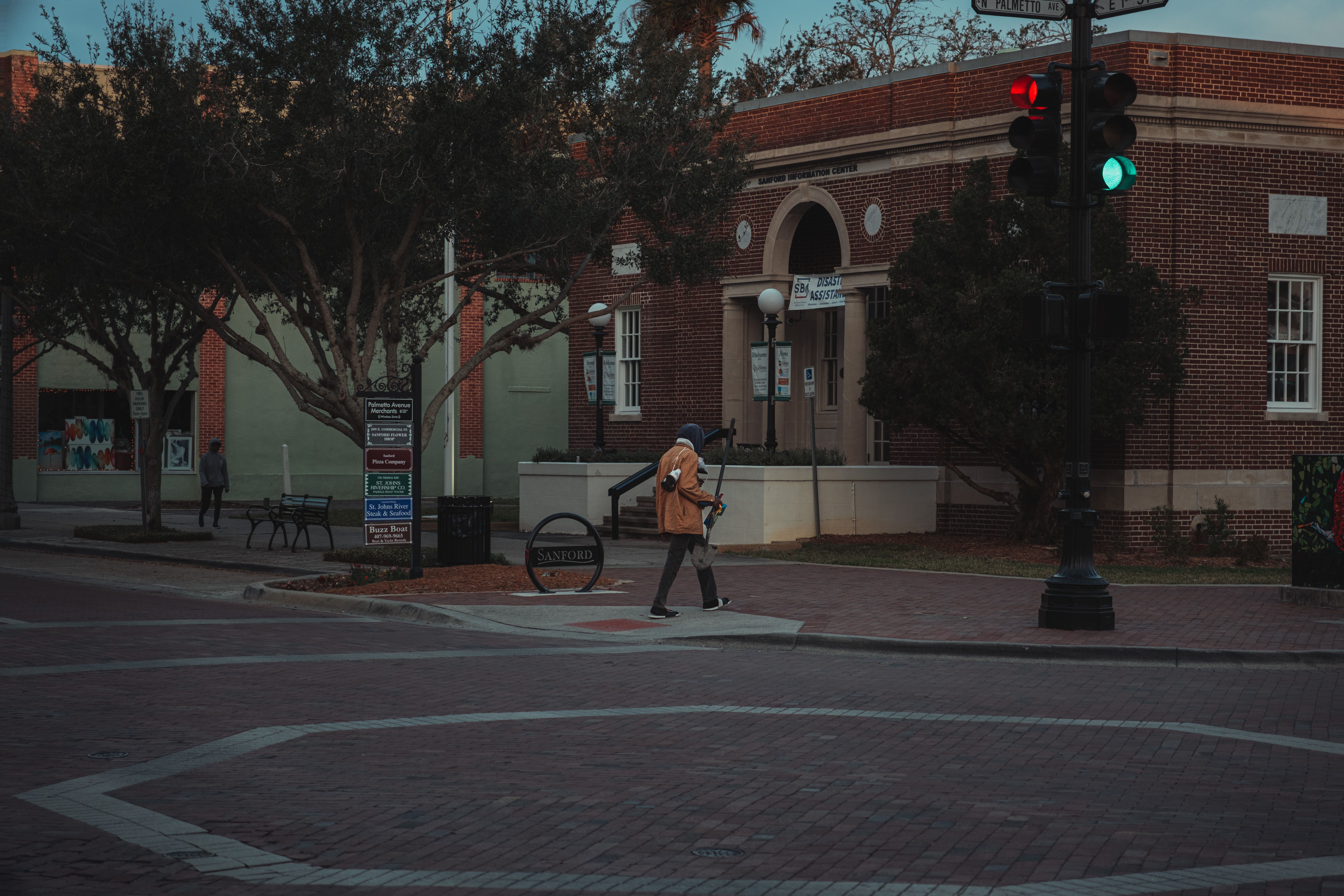 A person walking through an urban street intersection at twilight near brick buildings and trees.