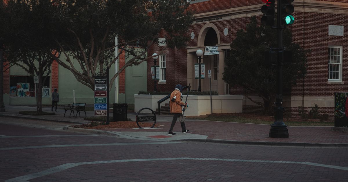 A person walking through an urban street intersection at twilight near brick buildings and trees.