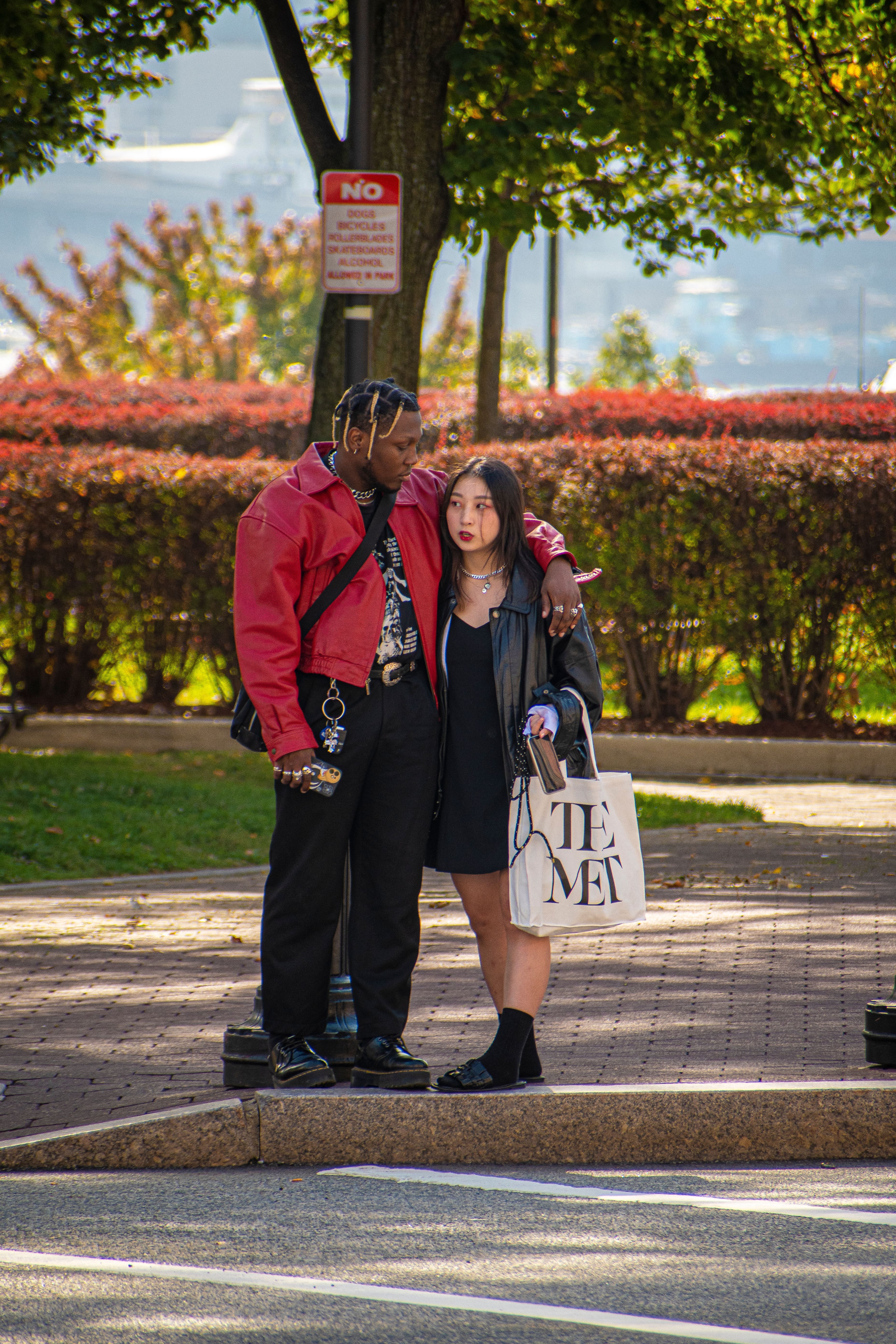A couple embracing on a sidewalk with greenery and cityscape in the background.