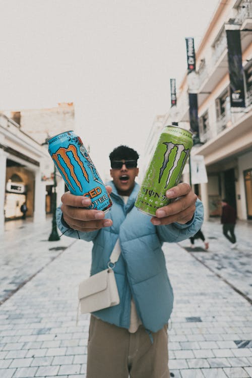 Man in a Blue Jacket Standing on a Street and Holding Two Cans of Monster Energy Drinks