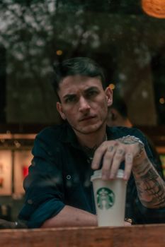 A tattooed man holding a coffee cup, seen through a window with a moody ambiance.