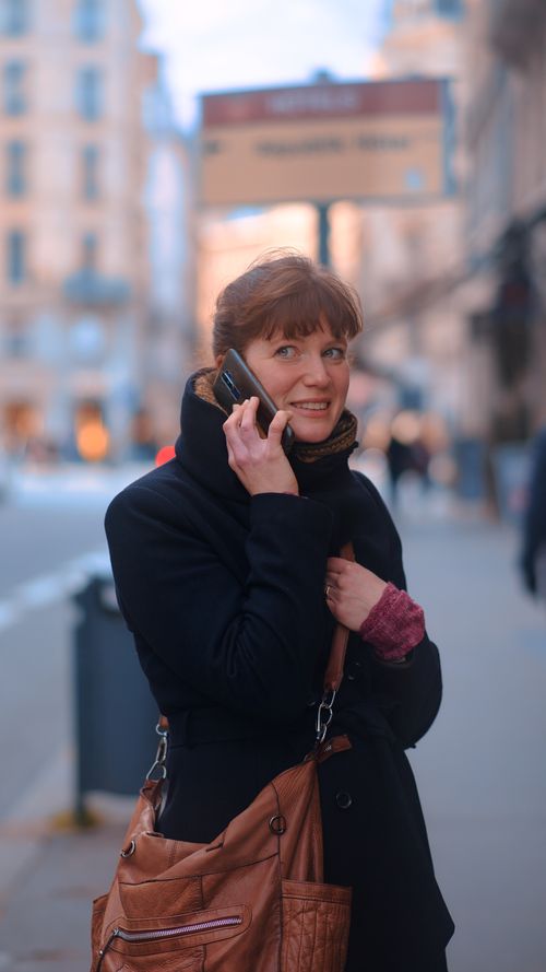 Free Smiling woman on phone in downtown area, wearing a coat and carrying a bag. Stock Photo